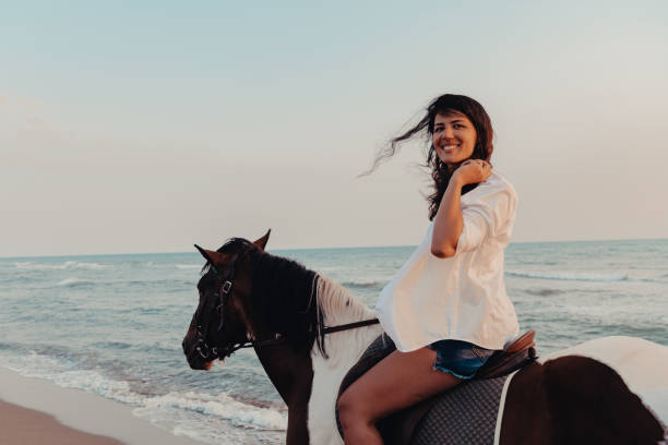 kobieta w letnich ubraniach lubi jeździć konno po pięknej piaszczystej plaży o zachodzie słońca. selektywne ustawianie ostrości - cowboy horseback riding nature blue zdjęcia i obrazy z banku zdjęć