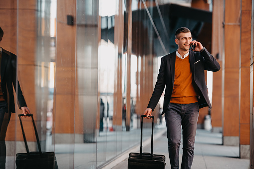 Going to airport terminal. Confident businessman traveler walking on city streets and pulling his suitcase drinking coffee and speaking on smartphone. High quality photo