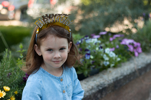 A little beautiful preschooler girl with a crown happy birthday on her head stands in a denim blue dress against the background of flowers
