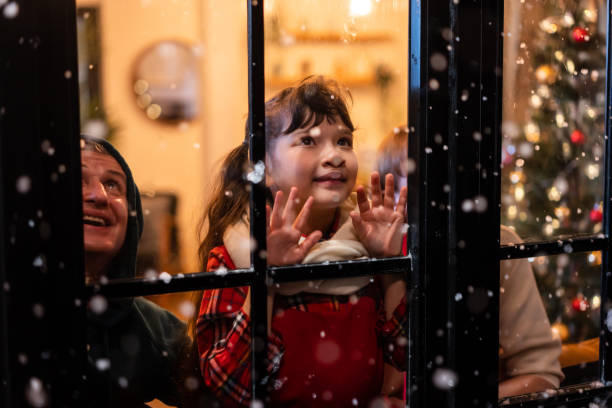 adorable niño mira a través de la ventana y admira los primeros copos de nieve. niña sentada en la sala de estar de su casa, viendo la nieve caer afuera disfrutar de celebrar la navidad, acción de gracias con la familia. - christmas window santa claus lighting equipment fotografías e imágenes de stock