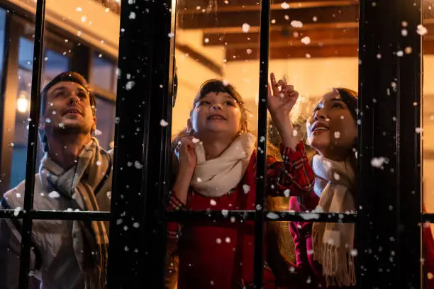 Photo of Adorable child looking at the window and first snow flakes with family. Young kid girl sitting in living room at home, watch snow falling outside enjoy celebrate Christmas, Thanksgiving with parents.