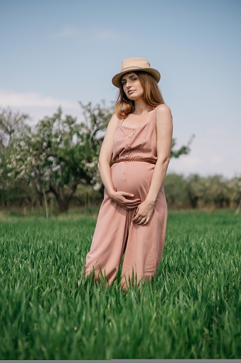 beautiful pregnant girl in overalls with a straw hat on a green field on a sunny spring day. High quality photo