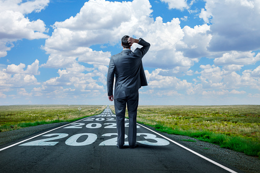 A businessman stands on and looks down a long, straight, rural road that has 2023 and subsequent future years painted on it. In the distance, as the road meets the horizon, puffy clouds punctuate the blue sky.