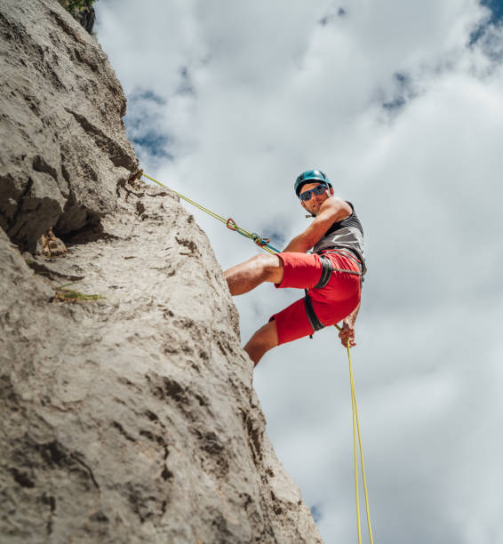 Active climber middle age man in protective helmet looking at camera while abseiling from cliff rock wall using rope with belay device and climbing harness. Active extreme sports time spending concept escalador activo hombre de mediana edad con casco protector mirando a la cámara mientras hace rappel desde la pared de roca del acantilado usando una cuerda con dispositivo de aseguramiento y arnés de escalada. concepto de gasto de tiempo en deportes ext