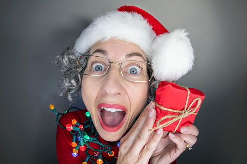 A funny fisheye image of a middle aged woman in a Santa hat excited about the small present she is holding.
