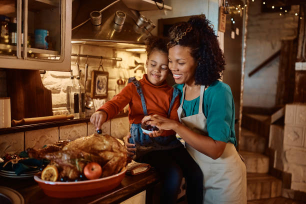 Happy black mother and her small daughter decorating roast turkey for Thanksgiving lunch. Happy African American mother and daughter enjoying preparing Thanksgiving turkey for family dinner in the kitchen. family dinners and cooking stock pictures, royalty-free photos & images
