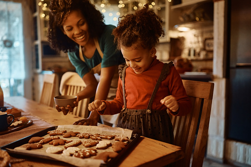 Happy African American mother and daughter enjoying in making gingerbread cookies for holidays in the kitchen. Focus is on girl.