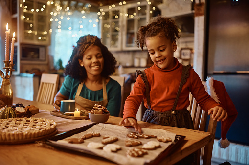 Happy African American little girl and her mother enjoying while making cookies on Christmas day in the kitchen.