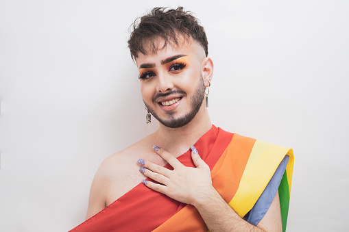 A young man in makeup posing against a white background with an LGBT flag while smiling and holding a hand on his chest.Diversity concept.