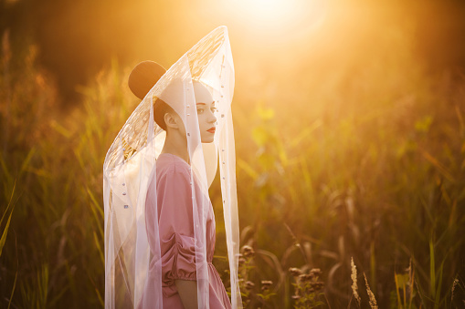 Geisha in kimono. Asian woman in hat with red lips on background of high green grass. Beauty makeup. Asian fashion. Sunset light