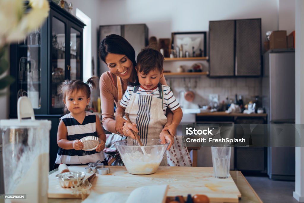 Family Making A Cake Together In The Kitchen Caucasian mother and her two little children making a cake from the ingredients. Cooking Stock Photo