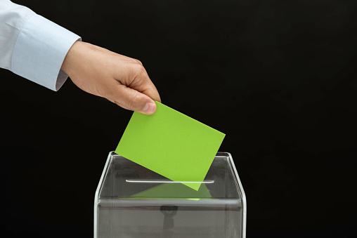 Human hand is inserting green envelope into ballot box in front of black background. Representing sustainable lifestyle.