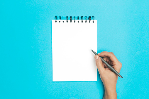 Hand is holding pencil and is about to write something into a spiral notepad on blue background.