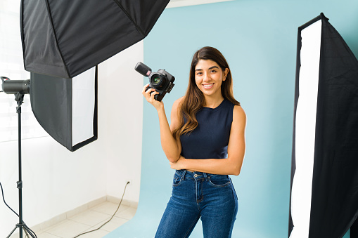 Happy beautiful woman photographer holding her professional camera and ready to take pictures at the studio
