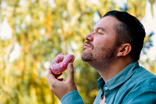 Hispanic handsome Man eating pink glazed donut outdoor in the park. Adult male enjoying in sweet treat dessert. Side view, head shot, copy space