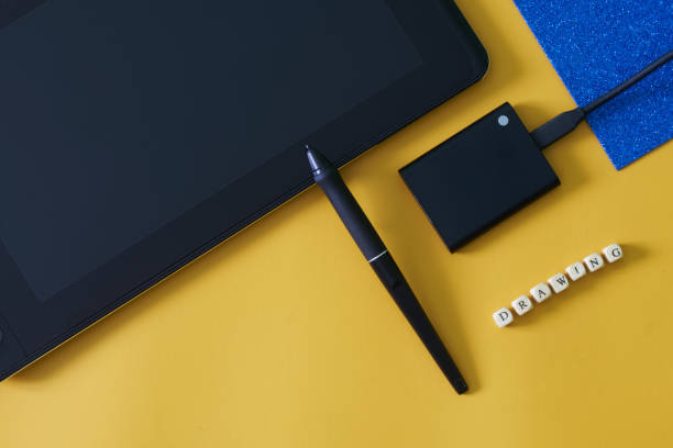 istock Graphic tablet with an interactive screen and a stylus on a yellow-blue geometric background next to an external ssd and the inscription drawing. Modern creativity and computer technologies. 1435449324