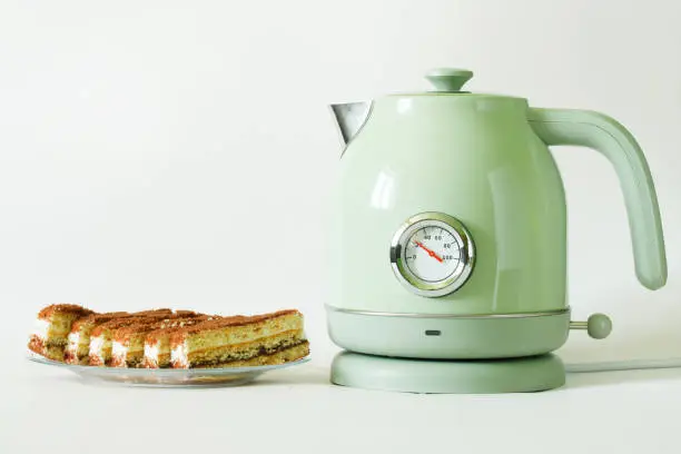 Trendy vintage-style electric kettle with a thermometer stands on a rotating stand, next to cakes. White background. Copy space. Close-up