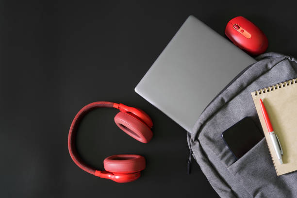 istock Gray textile urban backpack with laptop, mobile phone, pen and notepad on black background next to red headphones and mouse. Study, travel, trip and lifestyle with gadgets. Freelance tools. 1435449479