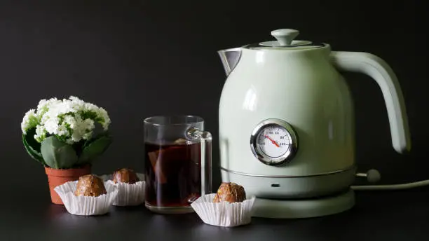 Trendy, vintage-style electric kettle with a thermometer stands next to a mug of tea, cakes and a small pot of white spring flowers. Black background. Copy space. Selective focus. Close-up