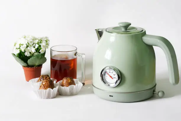 Trendy, vintage-style electric kettle with a thermometer next to cakes, a mug of tea, and a small pot of white spring flowers. White background. Copy space. Selective focus. Close-up