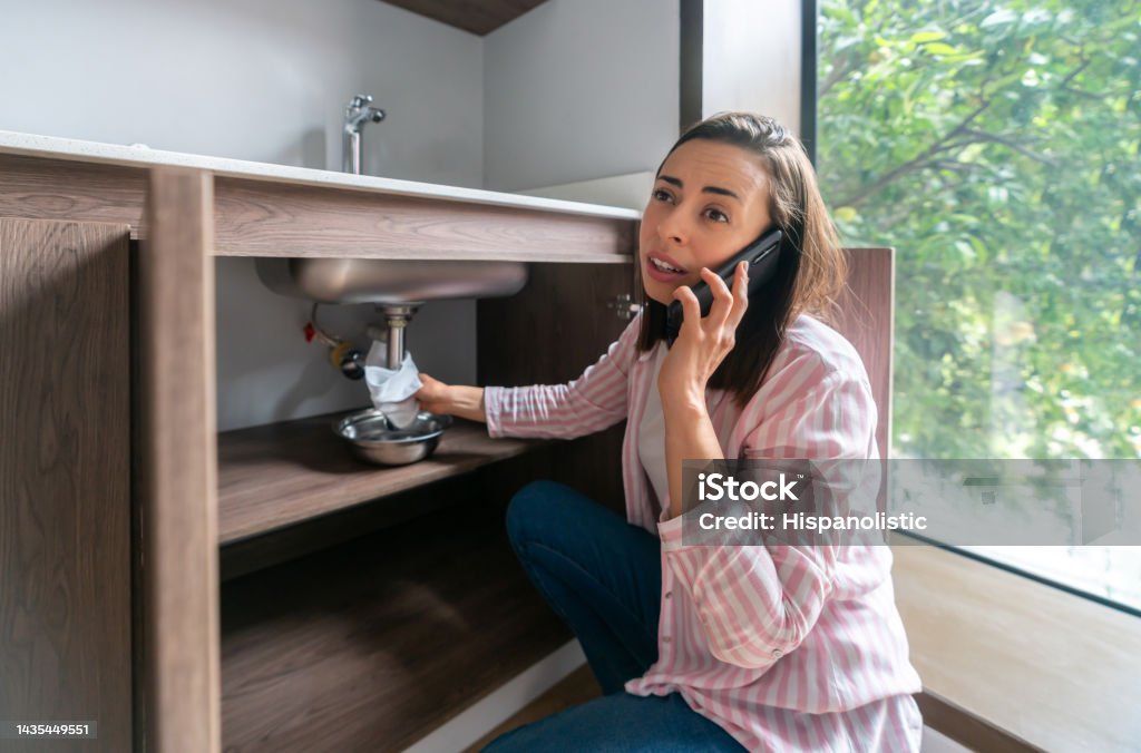 Woman having a leak in her kitchen sink and calling the plumber Latin American woman having a leak in her kitchen sink and calling the plumber Plumber Stock Photo