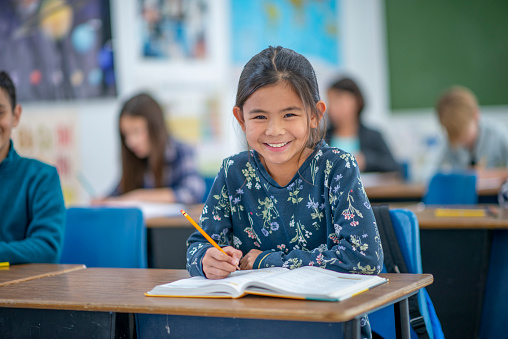 A young female student of Asian decent, sits at her desk as she works away on a writing assignment.  She is dressed casually and is looking up form her work to smile.