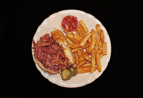 An open pastrami sandwich on a white, fancy plate.  Side of french fries and slices of pickle.  Ramiken of ketchup.   Black background.  View from directly above.