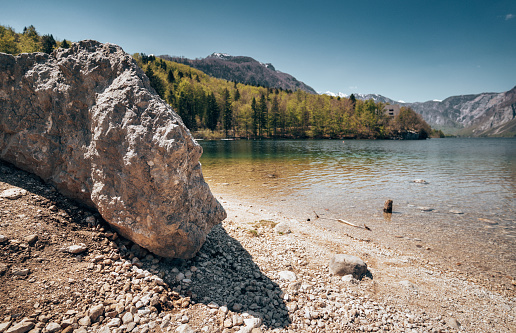 Rocks on the shoreline of a tranquil lake on a gloomy overcast day surrounded by forested hills and mountains reflected in the water