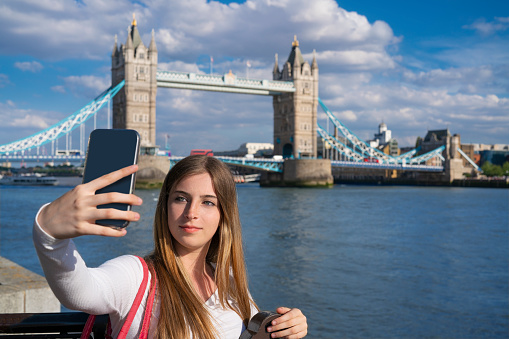 London tourist young blond woman shooting smartphone selfie photo in Tower Bridge over Thames river in a sunny blue sky summer day in UK, England. Great Britain, United Kingdom