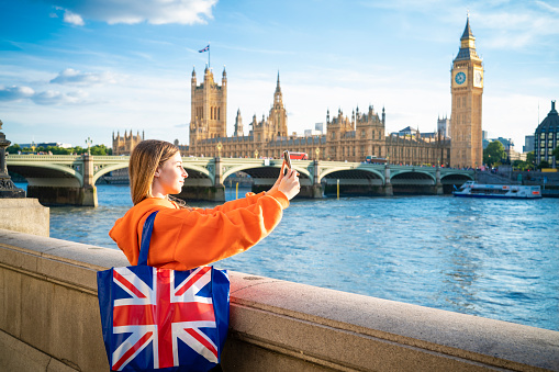 London blond tourist young student woman with UK flag shopping bag shoothing smartphone Great Britain United Kingdom, England at the evening dusk