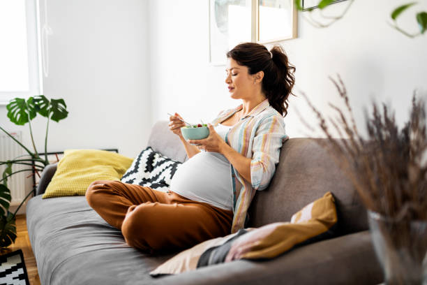 Young pregnant woman eating fresh salad sitting on a sofa stock photo