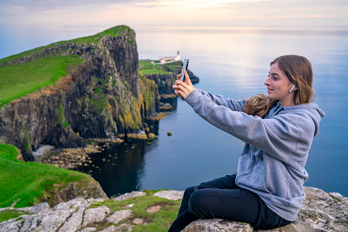 Blond girl shooting smartphone selfie photo in Skye island Nest Point lighthouse in Highlands Scotland UK in United Kingdom