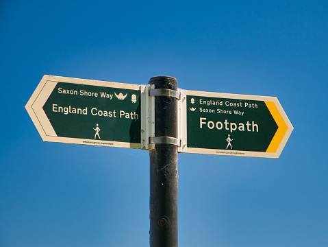 Two green signs on a grey metal post point the directions of the England Coast Path and Saxon Shore Way. Taken on a sunny day in summer with a clear, blue sky.