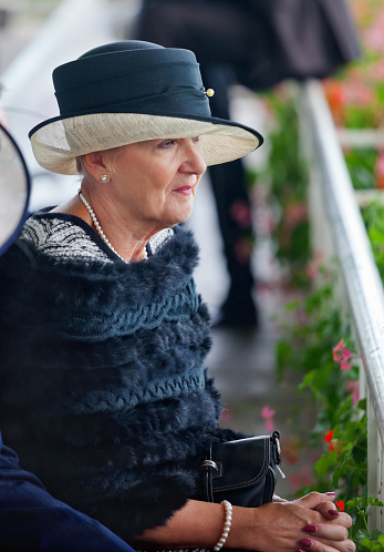 An elegant and stylish, mature lady dressed in a classic black and white outfit, with pearl necklace, earrings and bracelet seated in the racecourse grandstand at the horse racing on Ladies Day.