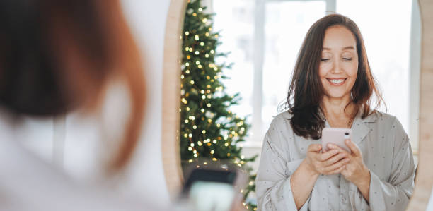 joven adulto de cuarenta años de mediana edad hermosa mujer con cabello oscuro en pijama acogedor mirando al espejo y tomándose selfie en la mañana de navidad en la habitación de la casa - 40 44 years fotografías e imágenes de stock