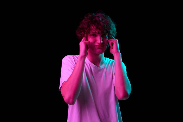 istock Terrible headache. Young man with discontent and undercurrent facial expression covering his ears isolated over dark background in neon light. 1435575094