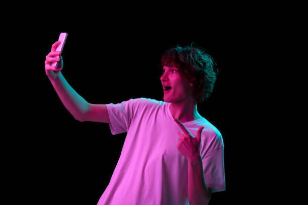 istock Taking selfie. Young handsome boy using smartphone isolated on blackbackground in neon light, filter. 1435575120