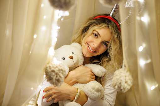 Close-up shot of beautiful young woman in pajamas and reindeer horns hugging teddy bear under baldachin in bedroom during Christmas holidays at cozy home