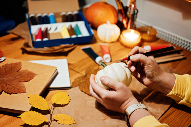 istock Autumn Thanksgiving pumpkins craft. DIY Fall Crafts for Adults. Female hands draw patterns on white pumpkin against autumn decor background on the table at home 1435577843