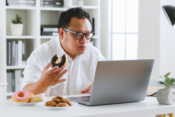 Asian fat man eating donut, sweet, junk food during working with computer laptop, unhealthy eating concept stock photo