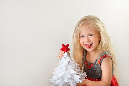 portrait of charming, cheerful girl in Christmas wreath, white dress showing heart sign with hands, isolated on white background with copy space.