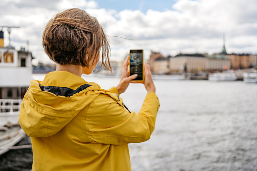 Beautiful young female tourist taking pictures of the view from the quayside using phone outdoors in Stockholm, Sweden.