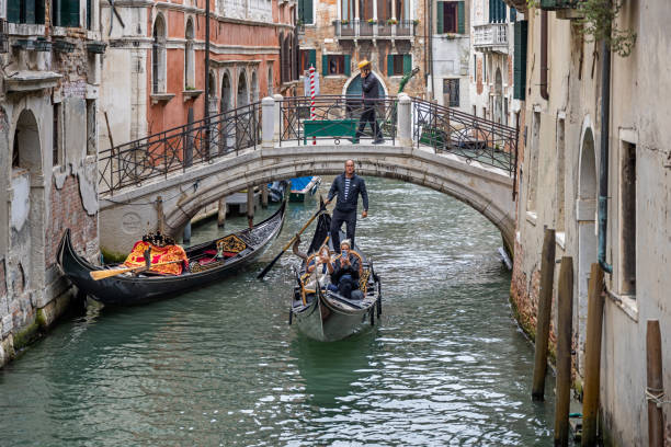 Venice, Italy - October 10th 2022: Gondolier with his gondola and two young female customers in a narrow canal in front of a bridge in the center of the old Venice