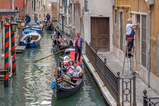 Venice, Italy - October 10th 2022: Everyday life with a row of gondolas filled with tourists in a narrow canal in the center of the old Venice while a skilled worker is standing on his ladder at the side