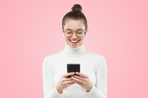 Young female in round glasses feeling relaxed and happy, laughing at funny content she is watching on smart phone screen, isoalated on pink background