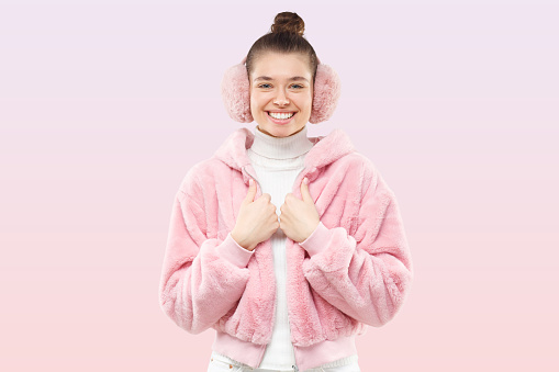Young female dressed in pink funky coat and white sweater, wearing ear warmers, smiling happily as if going to party, isolated on pink pastel background