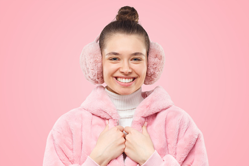 Close-up of young girl wearing bright pink fur coat and earmuffs, smiling happily, ready for photosession, isolated on pink background