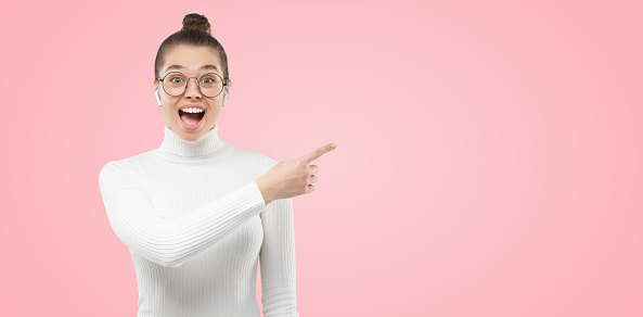 Banner of young excited female wearing eyeglasses and white turtleneck, pointing to copy space on right side, amazed by offer, isolated on pink background