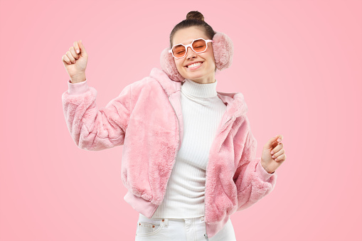 Young smiling woman wearing pink trendy fluffy bomber, ear warmers or winter headphones and trendy glasses, dancing isolated on pink background