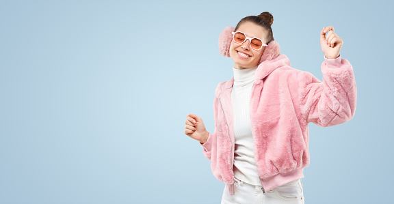 Horizontal banner of young female dancing in pink fluffy bomber, colored glasses and ear muffs, feeling happy at party, isolated on blue background